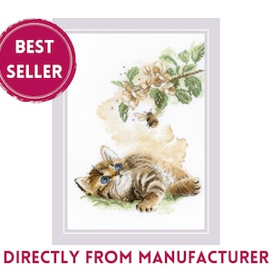 May include: Cross-stitch artwork featuring a tabby kitten with blue eyes, lying on green grass. A bee flies near a flowering branch. The image is framed and includes a "BEST SELLER" badge and the text "DIRECTLY FROM MANUFACTURER".