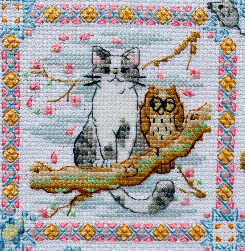 May include: A white cat with gray spots sits on a branch with a brown owl. The cat is looking to the right and the owl is looking to the left. The background is a light blue with pink flowers.