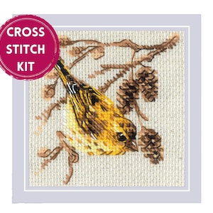 May include: Cross stitch kit featuring a detailed illustration of a yellow and brown bird perched on a branch with pinecones. The image is framed with a light gray border, and a pink circle with the text "CROSS STITCH KIT" is in the upper left corner.