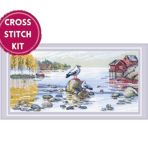 May include: Cross stitch kit featuring a scenic coastal landscape with two seagulls perched on a rock in the water. The scene includes red buildings, trees, and a serene body of water. The kit is framed in a light gray border.