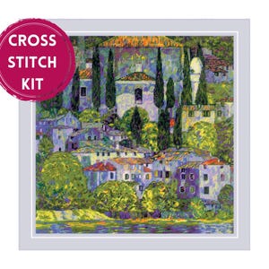 Peut inclure: Un kit de point de croix représentant une peinture paysagère impressionniste vibrante d'un village à flanc de colline avec des maisons en pierre, des cyprès et un feuillage vert luxuriant surplombant l'eau, avec le texte CROSS STITCH KIT en cercle.