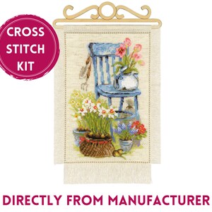 May include: Cross stitch kit featuring a detailed floral scene with a blue chair, potted flowers, and gardening tools. The kit includes a wooden hanger and a fringed fabric panel. The text "CROSS STITCH KIT" is in a pink circle.
