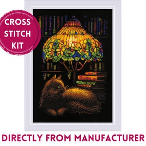 Op de afbeelding: Kruissteekpakket met een gezellige scène met een kat, boeken en een gebrandschilderde lamp. De lamp is verlicht en werpt een warme gloed op de kat en de boeken. De tekst "CROSS STITCH KIT" staat in een roze cirkel.