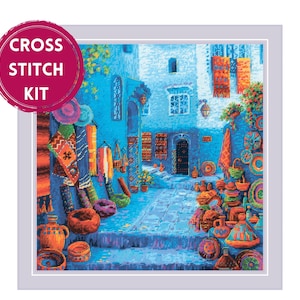 Puede incluir: Kit de punto de cruz que presenta una vibrante escena callejera con edificios azules, textiles coloridos y cerámica. La imagen incluye el texto "CROSS STITCH KIT" en un círculo rosa.