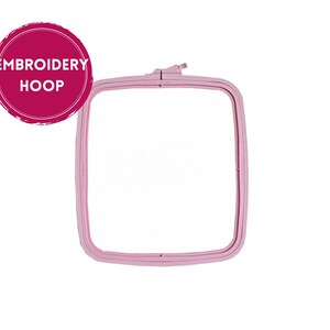 May include: A square, light pink embroidery hoop. The hoop is made of plastic and has a tightening mechanism at the top. A circular sticker with the words "EMBROIDERY HOOP" in white text is in the upper left corner.