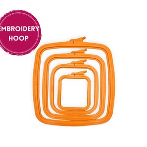 Op de afbeelding: Een stapel van vijf oranje vierkante borduurringen van verschillende maten. De ringen zijn gemaakt van plastic en hebben een sluiting aan de bovenkant. Een roze cirkel met de woorden "EMBROIDERY HOOP" staat in de linkerbovenhoek.