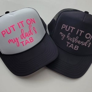 May include: Two black trucker hats with mesh back panels. One hat has white front panel with pink text that reads "PUT IT ON my dad's TAB". The other hat has a black front panel with pink glitter text that reads "PUT IT ON my husband's TAB".