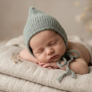 May include: A sleeping newborn wearing a sage green knitted bonnet with tie fastenings, resting on a stack of neutral-toned blankets. The bonnet has a pointed top and a textured knit pattern. The baby is asleep.