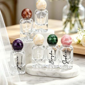 May include: A collection of small, clear glass perfume bottles with decorative, marble-like stoppers in various colors. Each bottle is labeled with a name and "Maid of honor" in elegant script. The bottles are arranged on a white marble surface.