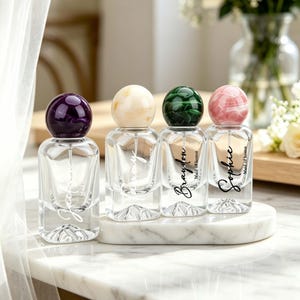 May include: Four small, clear glass perfume bottles with decorative, colorful spherical stoppers. Each bottle has a different name in cursive script: Josie, Tammy, Braydon, and Sophie. The bottles are on a white marble tray.