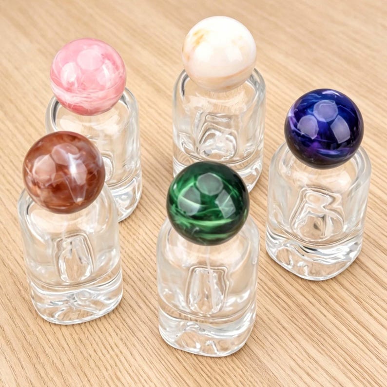 May include: Five small, clear glass bottles topped with decorative, colorful spheres. The spheres are in shades of pink, white, brown, green, and blue. The bottles are arranged on a light-colored wooden surface.