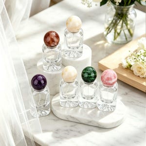 May include: Clear glass perfume bottles with colorful, spherical stoppers. The stoppers are in shades of brown, cream, purple, green, and pink. The bottles are arranged on white marble platforms, with a soft, natural light.
