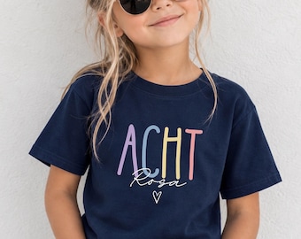 Könnte beinhalten: Ein dunkelblaues T-Shirt mit kurzen Ärmeln, das das Wort ACHT in großen, bunten Pastellbuchstaben zeigt, darunter das Wort Rosa in weißer Schreibschrift und ein kleines weißes Herz-Symbol unter dem Text.