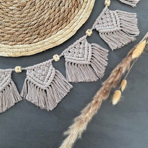 May include: A macrame banner in a neutral beige color, with wooden letter beads spelling "SIN". The banner has a geometric design with fringed edges. A woven placemat and dried ornamental grass are also in the image.