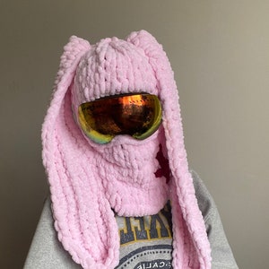 May include: Pink knitted bunny ear hat with a pair of ski goggles. The hat is made of a soft, fluffy yarn and has long, floppy ears.