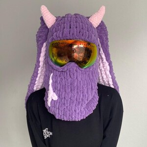 Puede incluir: M&aacute;scara de esqu&iacute; de punto morado y rosa con un par de gafas de esqu&iacute; naranjas. La m&aacute;scara tiene dos cuernos rosas en la parte superior.