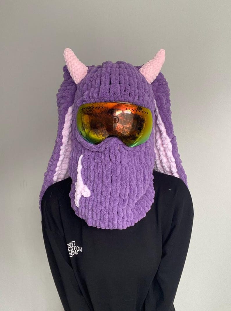 Puede incluir: M&aacute;scara de esqu&iacute; de punto morado y rosa con un par de gafas de esqu&iacute; naranjas. La m&aacute;scara tiene dos cuernos rosas en la parte superior.