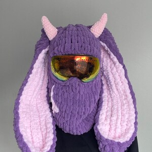 Puede incluir: M&aacute;scara de esqu&iacute; de punto morado y rosa con orejas de conejo y gafas de esqu&iacute;.