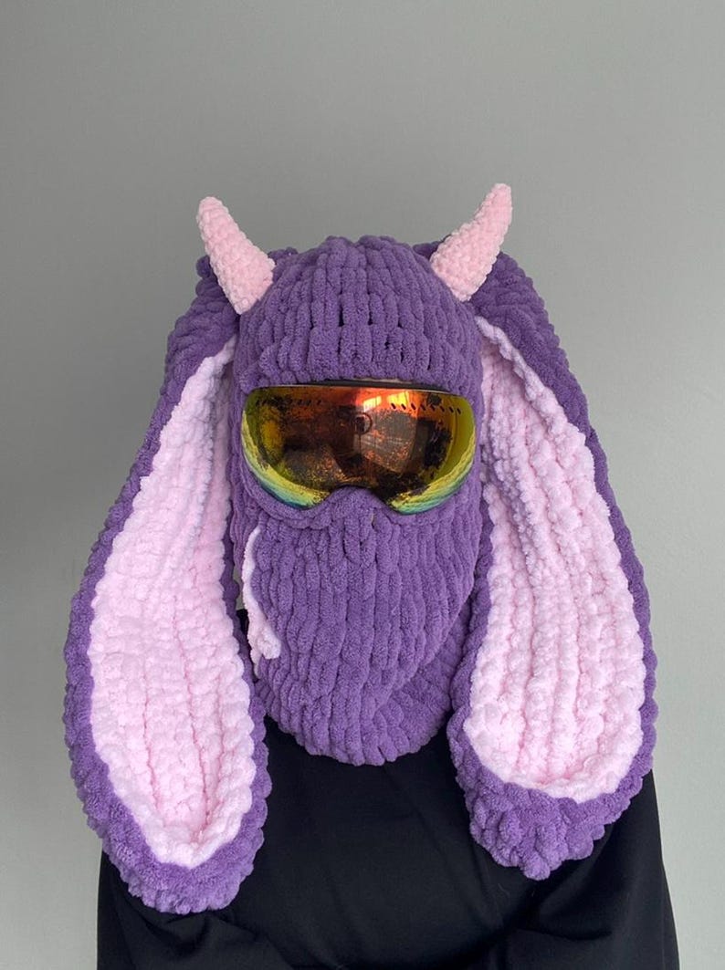 Puede incluir: M&aacute;scara de esqu&iacute; de punto morado y rosa con orejas de conejo y gafas de esqu&iacute;.