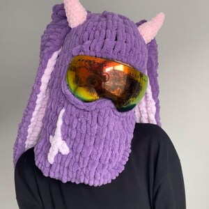 Puede incluir: M&aacute;scara de esqu&iacute; de punto morado con cuernos rosas y un par de gafas de esqu&iacute;. La m&aacute;scara tiene un dise&ntilde;o blanco en la parte delantera.