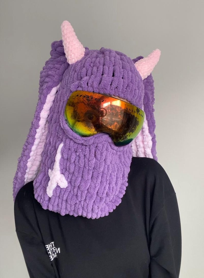 Puede incluir: M&aacute;scara de esqu&iacute; de punto morado con cuernos rosas y un par de gafas de esqu&iacute;. La m&aacute;scara tiene un dise&ntilde;o blanco en la parte delantera.