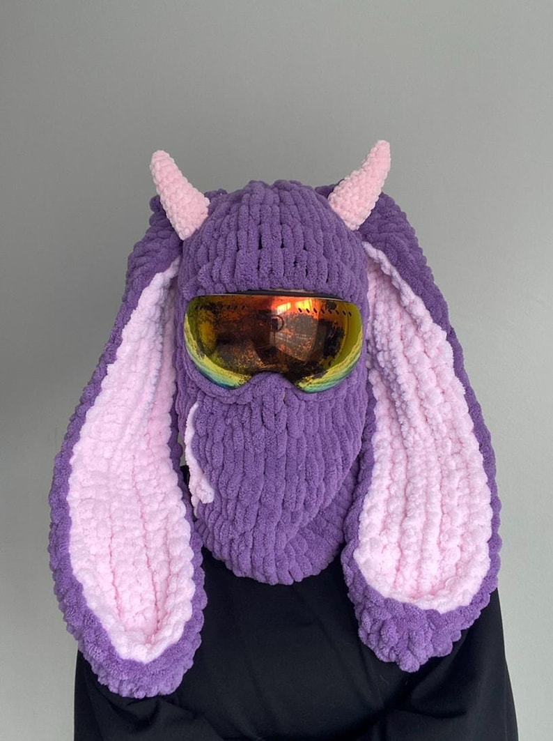 Puede incluir: M&aacute;scara de esqu&iacute; de punto morado y rosa con orejas de conejo y gafas de esqu&iacute;.