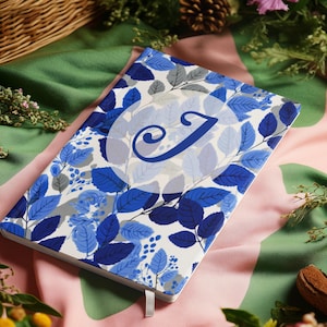 Könnte beinhalten: Ein Notizbuch mit blauem und weißem floralem Blattmuster. Das Cover zeigt einen großen blauen Buchstaben "J" in einem weißen Kreis. Ein weißes Band-Lesezeichen ist sichtbar.