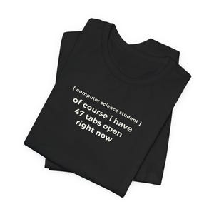 May include: Black t-shirt with white text: "[computer science student] of course i have 47 tabs open right now." The shirt is folded and appears to be made of soft material. A humorous design for computer science students.