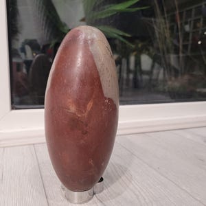 May include: A brown and grey stone lingam, a sacred object in Hinduism, is displayed on a silver stand. The lingam is oval-shaped and has a smooth, polished surface.