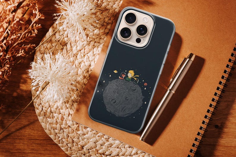 Puede incluir: Funda de tel&eacute;fono azul oscuro con una ilustraci&oacute;n de El Principito en un planeta. Un bol&iacute;grafo dorado y un cuaderno est&aacute;n junto a la funda sobre una superficie tejida. Flores blancas y plantas secas en el fondo.