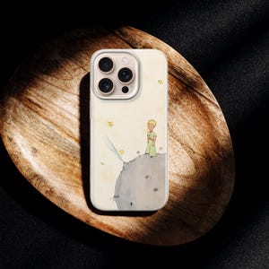Puede incluir: Una funda de tel&eacute;fono con una ilustraci&oacute;n en acuarela de un personaje de "El Principito" en un planeta. La funda es blanquecina con un borde transparente y un recorte para la c&aacute;mara multiobjetivo. La funda est&aacute; sobre una bandeja de madera.