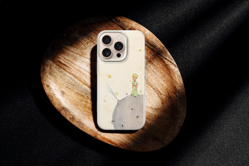 Puede incluir: Una funda de tel&eacute;fono con una ilustraci&oacute;n en acuarela de un personaje de "El Principito" en un planeta. La funda es blanquecina con un borde transparente y un recorte para la c&aacute;mara multiobjetivo. La funda est&aacute; sobre una bandeja de madera.