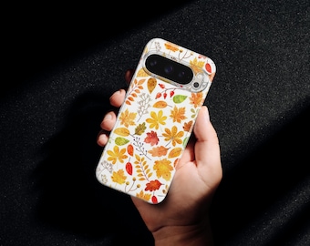May include: A white phone case featuring a colorful autumn leaf pattern. The design includes various leaves in shades of yellow, orange, and red, along with small berries and sprigs. The case is held in a hand against a dark background.