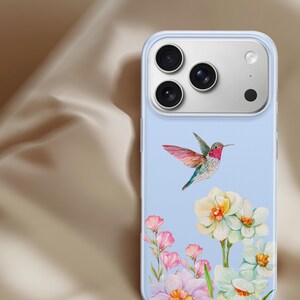 Op de afbeelding: Lichtblauwe telefoonhoes met een aquarelontwerp van een kolibrie in vlucht boven witte en roze bloemen. De hoes heeft een transparante rand en een cameramodule met drie lenzen.