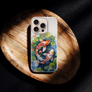 May include: A phone case featuring a watercolor painting of two koi fish swimming in a pond with lily pads. The fish are orange and white, set against a blue and green background. The case is on a wooden tray.