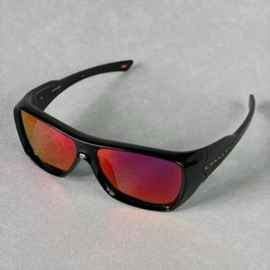May include: Black sunglasses featuring a wrap-around frame design and reflective red lenses. The temples display the Oakley brand name in a metallic finish. These sunglasses have a single-piece lens construction with a prominent bridge detail.