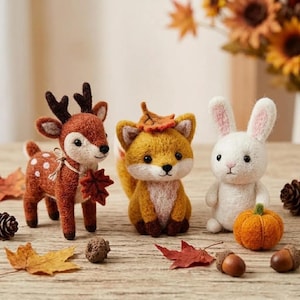 May include: Three needle-felted animal figurines: a brown deer with white spots and a leaf, a golden-orange fox with a leaf on its head, and a white rabbit next to a small orange pumpkin, all arranged on a wooden surface with scattered leaves.