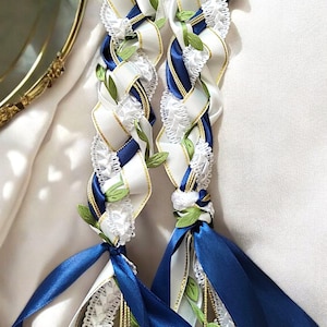 May include: Two braided leis made with white, blue, and gold ribbon, accented with green leaves and white flowers.
