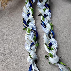 May include: Two white and blue braided ribbon leis with green leaves and white lace accents.