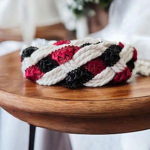 May include: A braided belt or accessory constructed from thick white rope interwoven with strips of black and deep red fabric. The design features a repeating pattern of white, black, and red segments, creating a textured, multi-colored appearance.