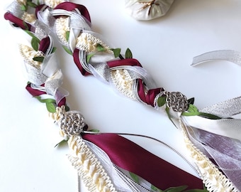 Peut inclure: Un ruban de cérémonie de mariage tressé blanc et bordeaux avec des accents argentés et ivoire. Le ruban est décoré de petites feuilles vertes et a un charme argenté à chaque extrémité. Le ruban est présenté avec deux colombes en porcelaine blanche.