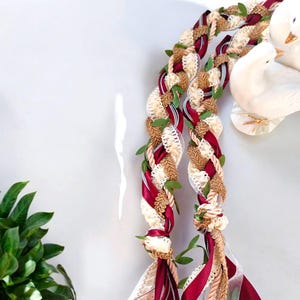 Puede incluir: Dos leis trenzados, uno en burdeos y otro en beige, con hojas verdes entrelazadas. Los leis están atados en la parte superior.