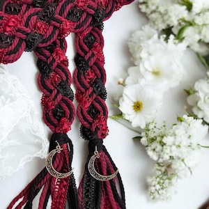 Op de afbeelding: Gevlochten haaraccessoires in rood en zwart, versierd met gouden halve maan bedels. De vlechten zijn ingeweven met linten en touw, wat een gotische of romantische uitstraling creëert. Witte bloemen op de achtergrond.