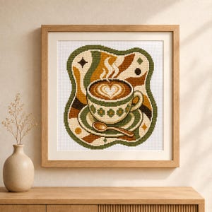 Könnte beinhalten: Ein gerahmtes Kreuzstich-Stickbild mit einer Kaffeetasse mit herzförmiger Latte-Art, einer Untertasse und einem Löffel. Das Design zeigt einen wellenförmigen grünen Rand mit abstrakten braunen, orangefarbenen und cremefarbenen Formen.