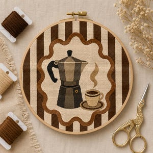 Puede incluir: Un bastidor de bordado circular de madera con un patrón de punto de cruz de una cafetera moka y una taza de café con vapor. El fondo presenta rayas verticales marrones y crema con un borde festoneado. Hilo de bordar y tijeras cerca.