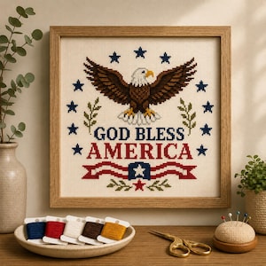 May include: A framed cross-stitch embroidery featuring a bald eagle with brown wings and a white head, surrounded by thirteen blue stars. The text GOD BLESS AMERICA is stitched in blue and red, accompanied by a red and white striped banner.