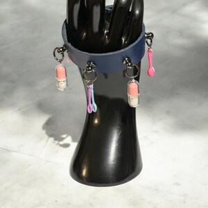 May include: A dark blue leather bracelet with multiple charms. The charms include two pink and white pill-shaped charms, a pink spoon charm, and a blue and purple spoon charm.