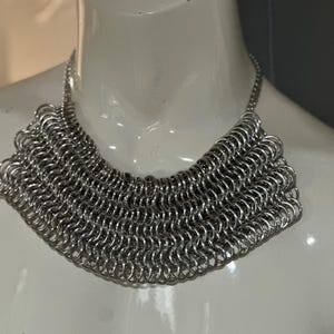 May include: A silver chainmail necklace with a wide, rounded collar design. The necklace is made of many small, interconnected metal rings.