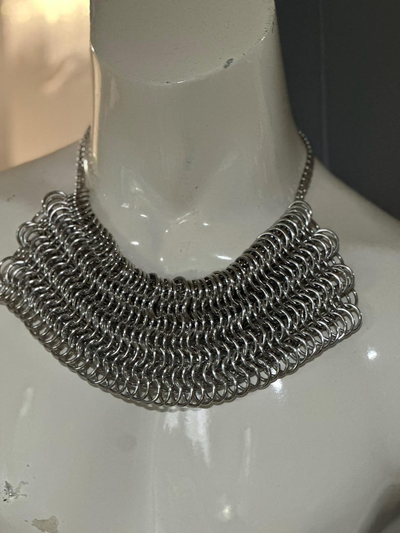 May include: A silver chainmail necklace with a wide, rounded collar design. The necklace is made of many small, interconnected metal rings.