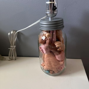 Puede incluir: Una lámpara de tarro de albañil con una tapa gris y un casquillo de luz plateado. El tarro está lleno de pequeñas piezas de muñeca de plástico, incluyendo brazos, piernas y cabezas.